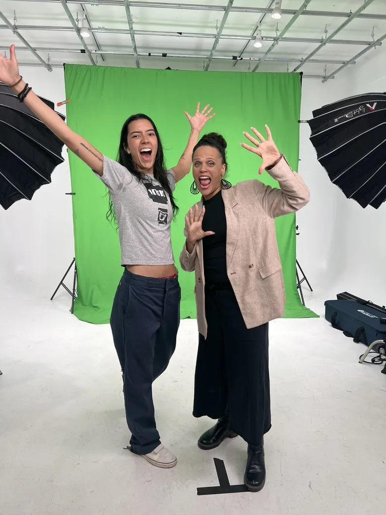 Rosa Lee Timm and a collaborator in front of a green screen, both with their arms raised and hands open, laughing.