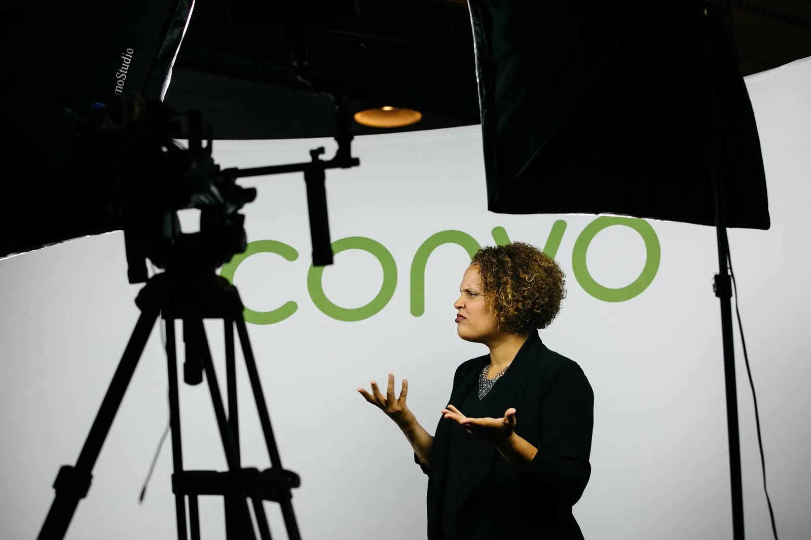 Rosa Lee Timm signing in a studio, lit by two large softboxes, a camera on a tripod in the foreground and a branded backdrop behind her.