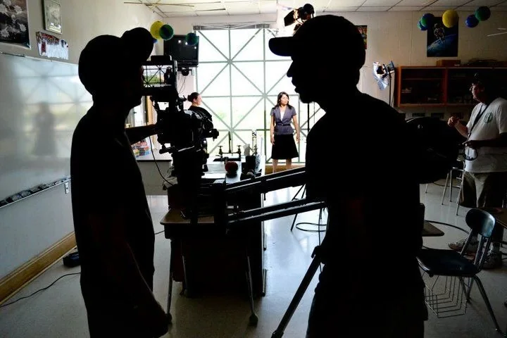 A camera crew silhouetted in the foreground of a classroom set, the performer lit in the distance beyond them.