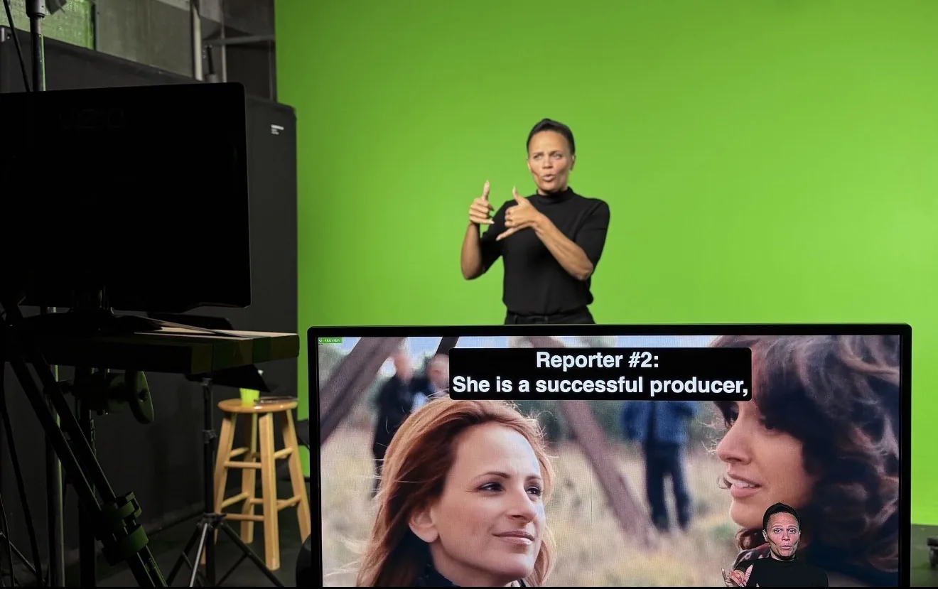 Rosa Lee Timm signing in front of a green screen in a studio. In the foreground, a production monitor shows the footage being worked on.