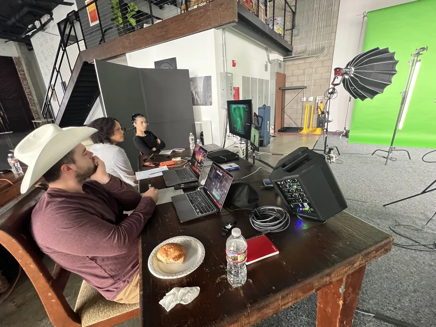 Rosa Lee Timm seated at a production table beside two colleagues, all watching a monitor. Laptops, a sound mixer, notes and coffee cover the table; a lit green screen stands at the edge of the room.