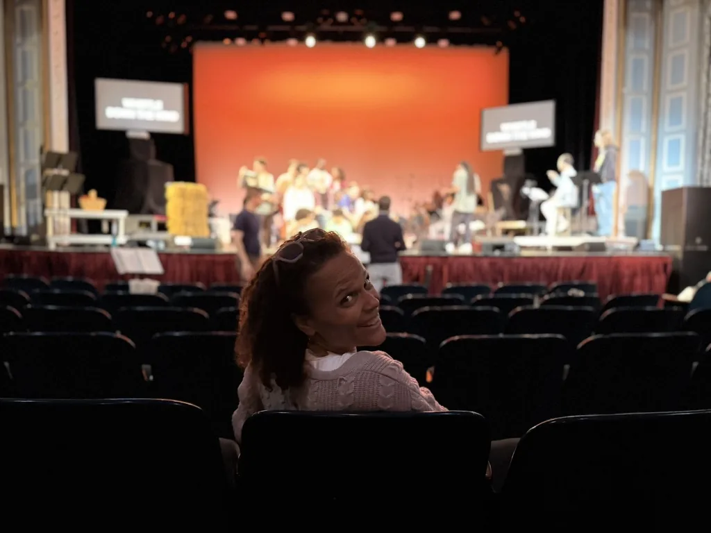 Rosa Lee Timm sitting in the empty seats of a theater, turned back toward the camera. Behind her, the company is on stage in rehearsal under warm light.
